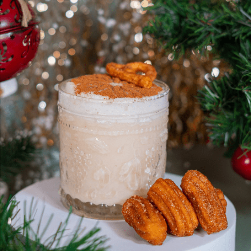 Churro Martini cremoso en vaso de vidrio con espuma, canela y mini churros sobre fondo navideño.