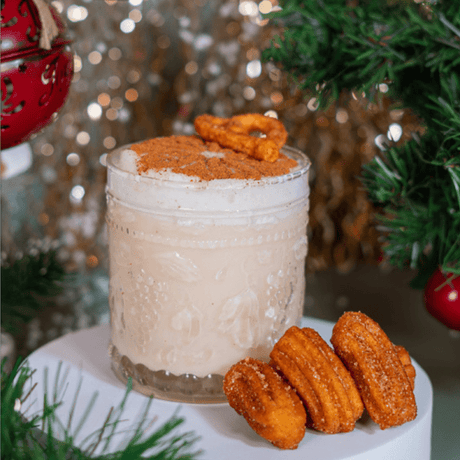 Churro Martini cremoso en vaso de vidrio con espuma, canela y mini churros sobre fondo navideño.