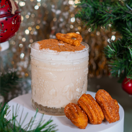 Churro Martini cremoso en vaso de vidrio con espuma, canela y mini churros sobre fondo navideño.